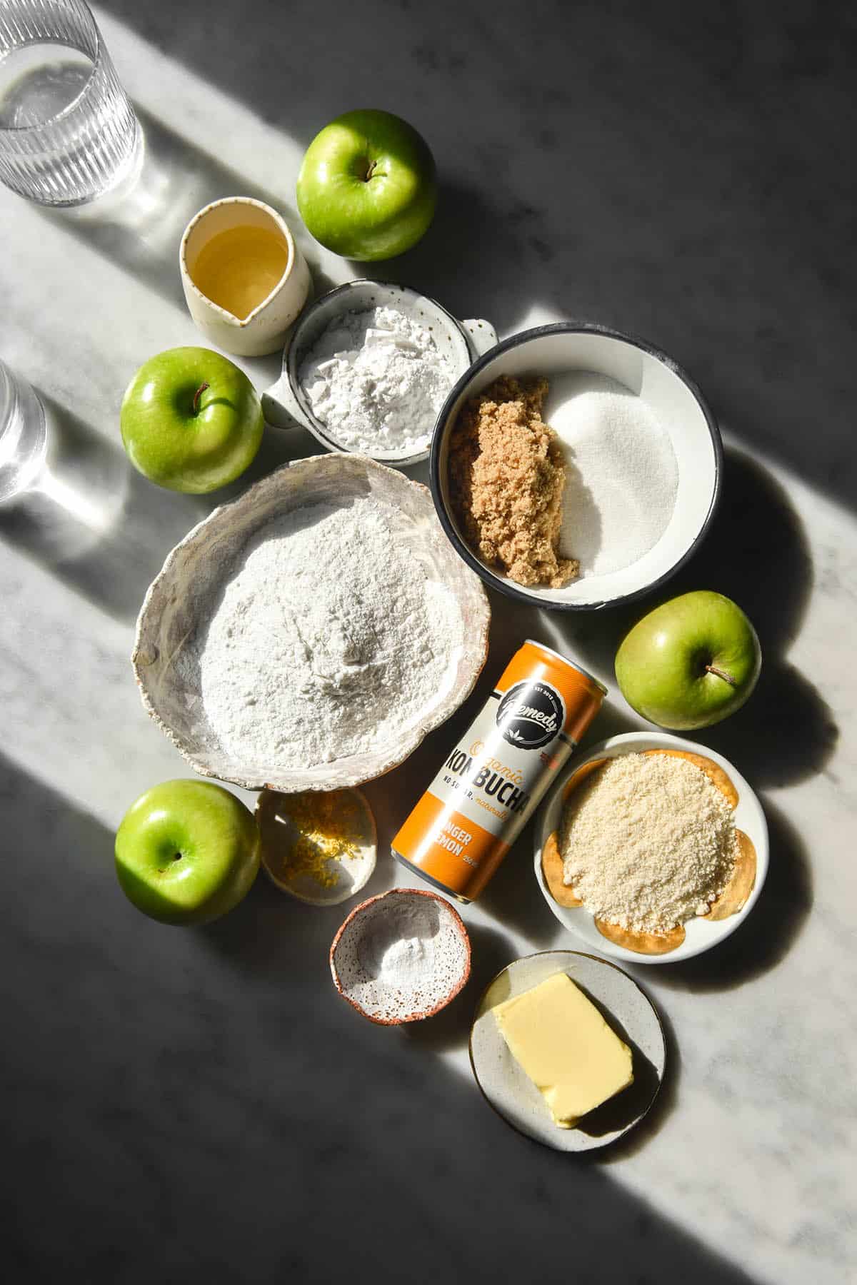 A sunlit view of the ingredients used for a gluten free apple cake in small white ceramic bowls atop a white marble table. The ingredients are surrounded by apples and sunlit water glasses sit to the left of the image