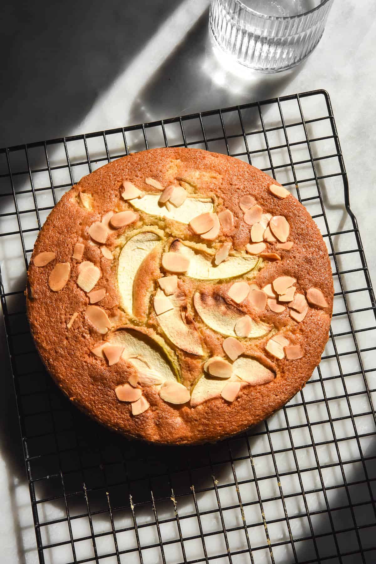 An aerial sunlit image of a gluten free apple cake on a wire cooling rack atop a white marble table. The cake is golden brown and topped with extra apple slices and sliced almonds. 