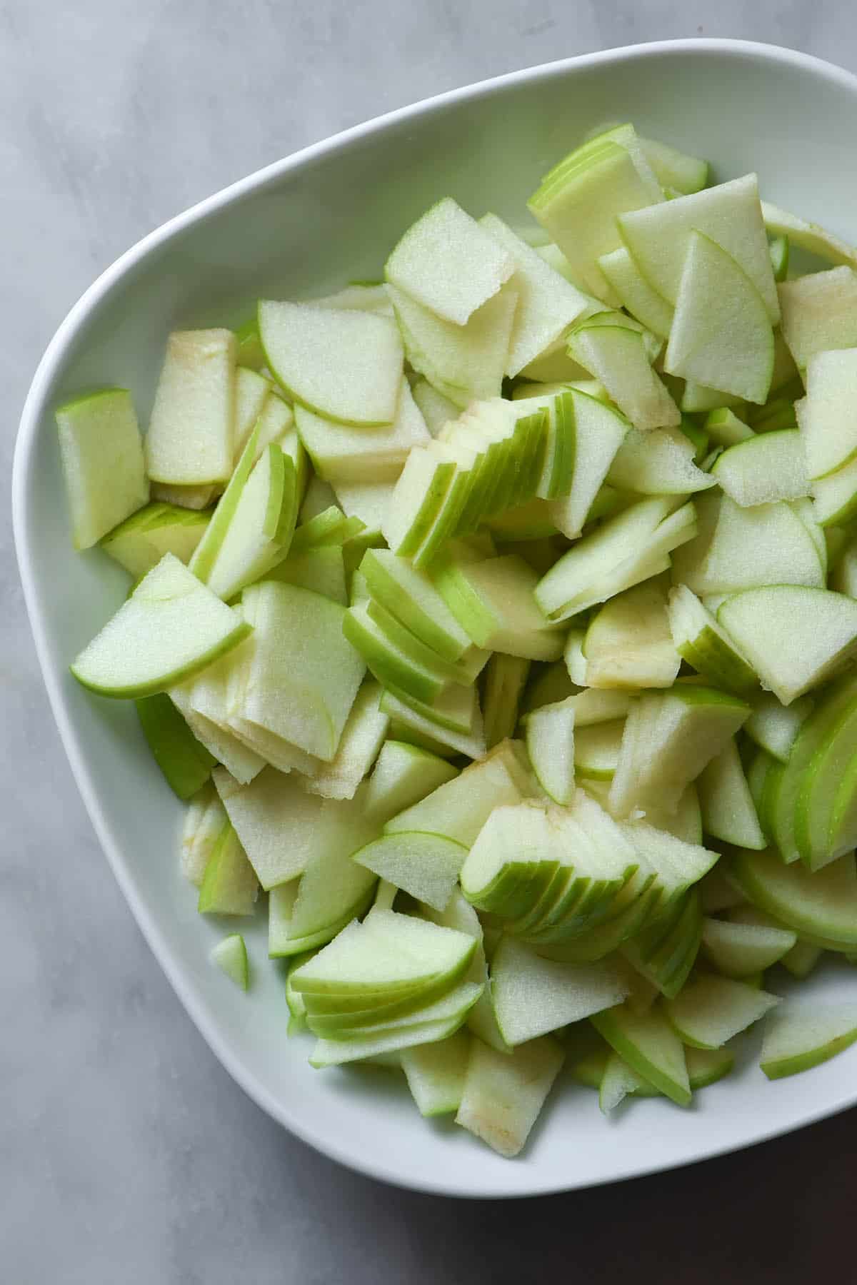 An aerial image of the slices of apple used for a gluten free apple cake. The slices sit in a white bowl on a white marble table. 