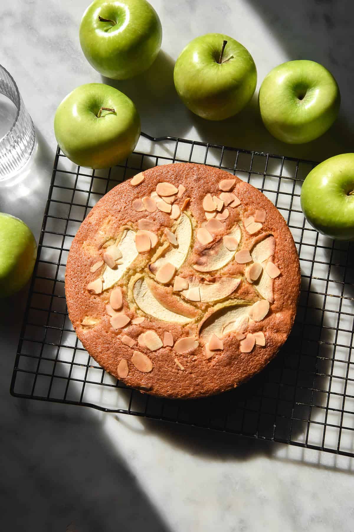 An aerial sunlit image of a gluten free apple cake on a wire cooling rack atop a white marble table. The cake is golden brown and topped with extra apple slices and sliced almonds. It is surrounded by green apples and a sunlit glass of water