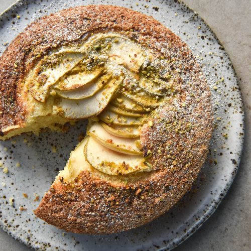 An aerial image of a gluten free apple cake on a white speckled ceramic plate. The cake sits on a light stone backdrop and a slice has been taken from the left side. It is topped with sliced apple, a sprinkle of crushed pistachio and icing sugar