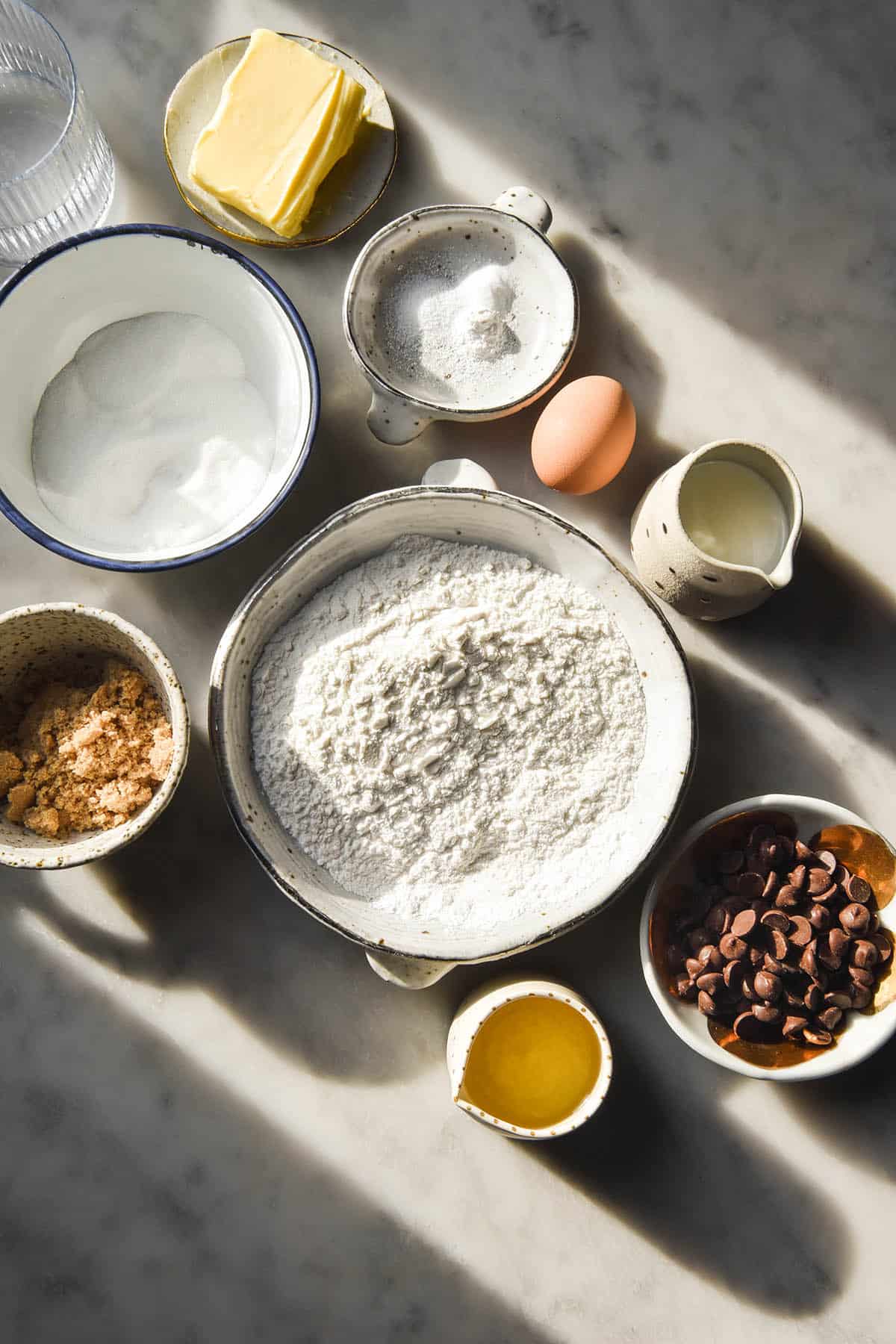 An aerial view of the ingredients needed to make gluten free chocolate chip muffins arranged atop a white marble table. The ingredients are in white bowls and in a patch of sunlight on the table