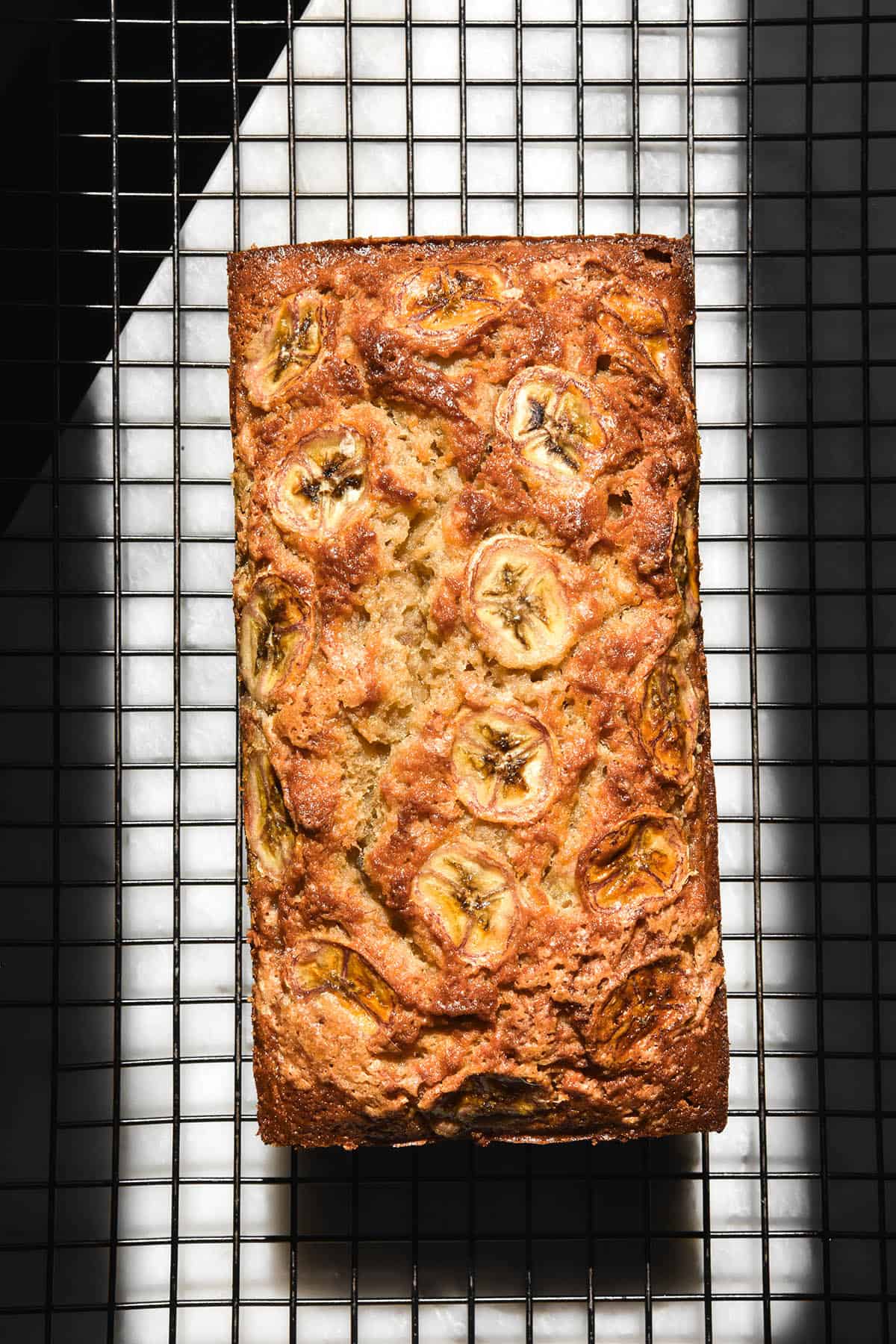 An aerial view of a gluten free dairy free banana bread loaf on a wire cooling rack atop a white marble table in contrasting sunlight