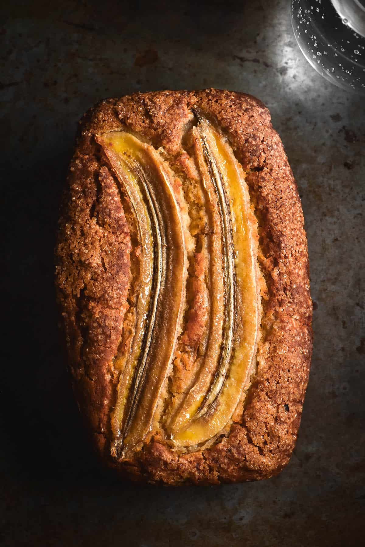 An aerial close up image of a loaf of gluten free dairy free banana bread. The banana bread sits on a dark steel backdrop. The banana loaf is golden brown and topped with slices of banana and finishing sugar