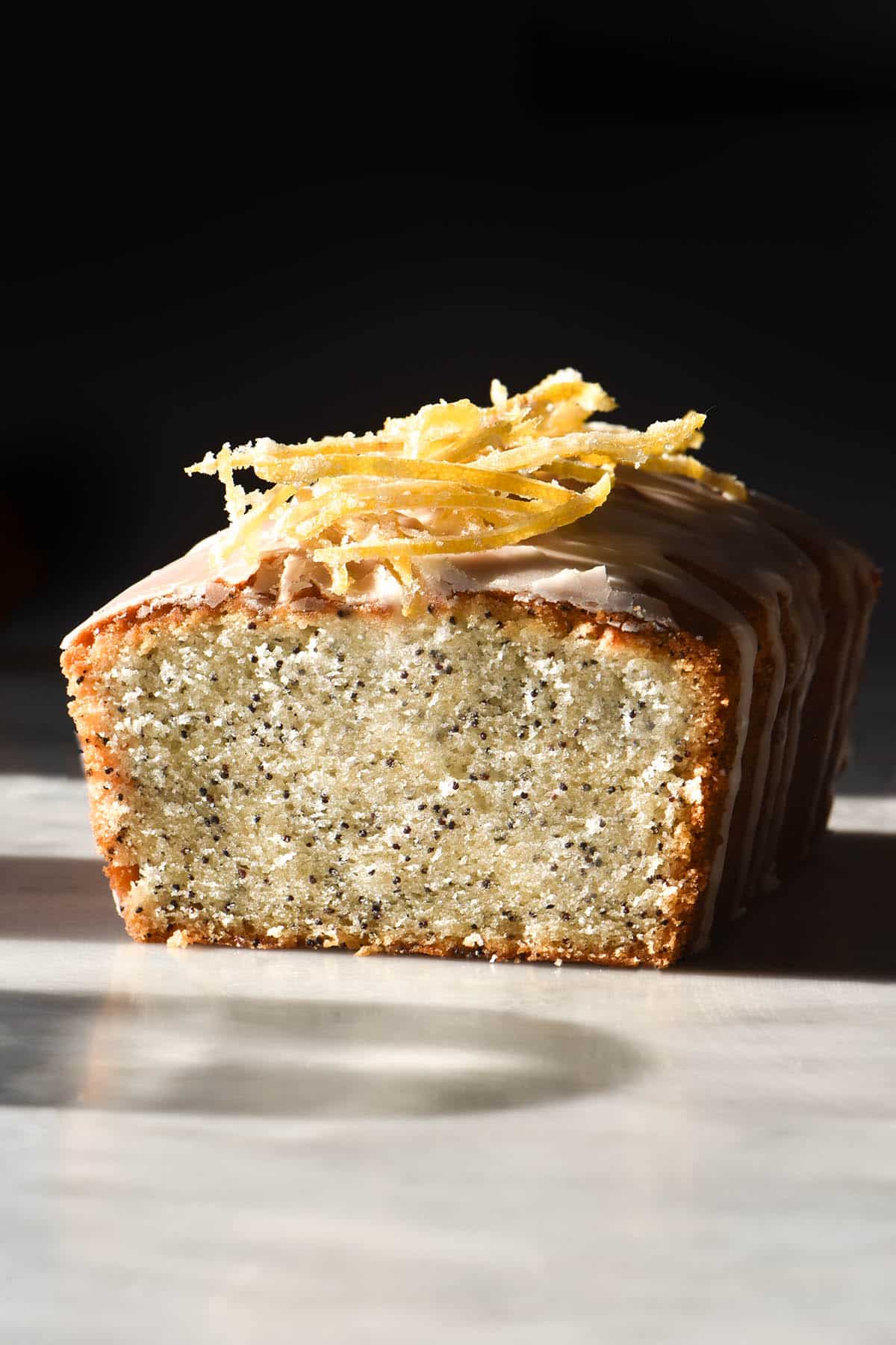 A side on view of a gluten free lemon poppyseed loaf topped with lemon icing and candied lemon. The loaf sits on a white marble table against a dark backdrop