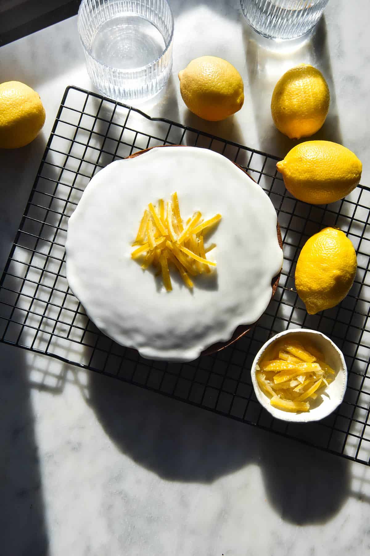 An aerial image of a gluten free lemon poppyseed cake in contrasting sunlight. The cake sits on a cake rack atop a white marble table. It is topped with lemon icing, candied lemon strips, and is surrounded by extra lemons and sunlit water glasses. 