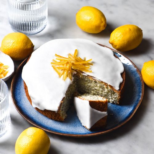 A side on image of a gluten free lemon poppyseed cake on a blue plate atop a white marble table. The cake is topped with lemon icing and candied lemon. It is surrounded by extra lemons and sunlit glasses of water.