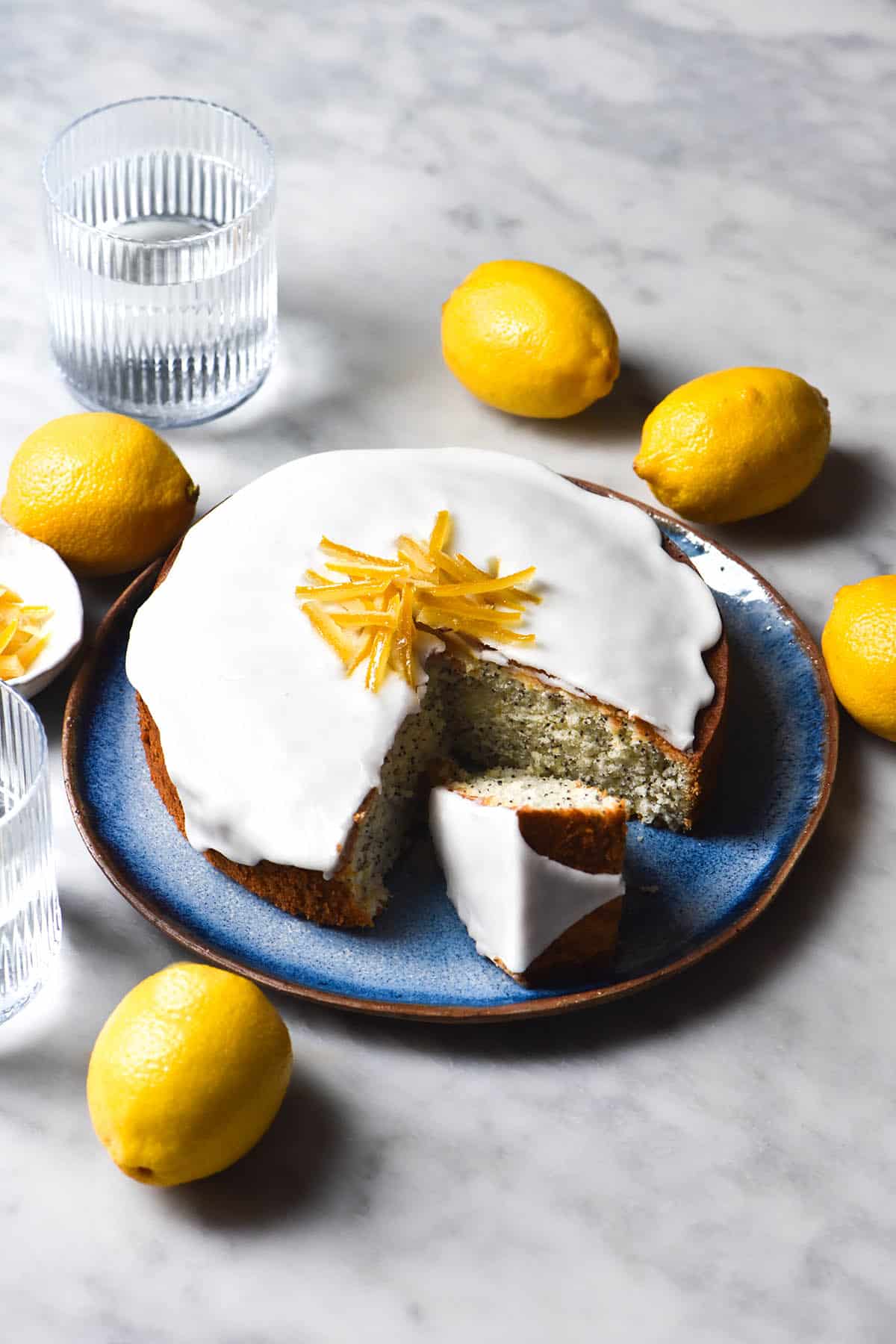 A side on image of a gluten free lemon poppyseed cake on a blue plate atop a white marble table. The cake is topped with lemon icing and candied lemon. It is surrounded by extra lemons and sunlit glasses of water. 
