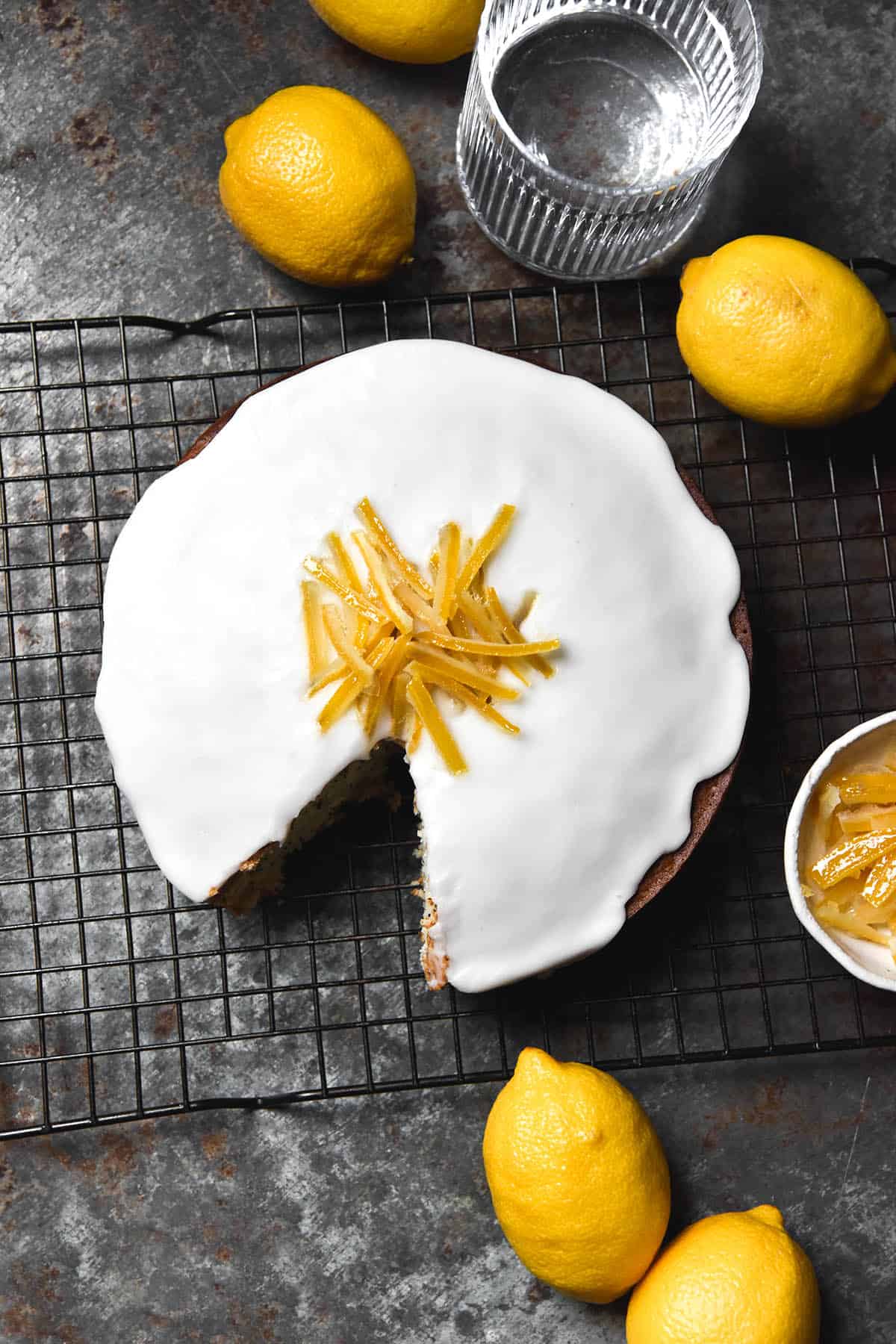 An aerial image of a gluten free lemon poppy seed cake sitting on a cake rack atop a dark steel backdrop. The cake is topped with lemon icing and candied lemons, and surrounded by extra lemons and water glasses