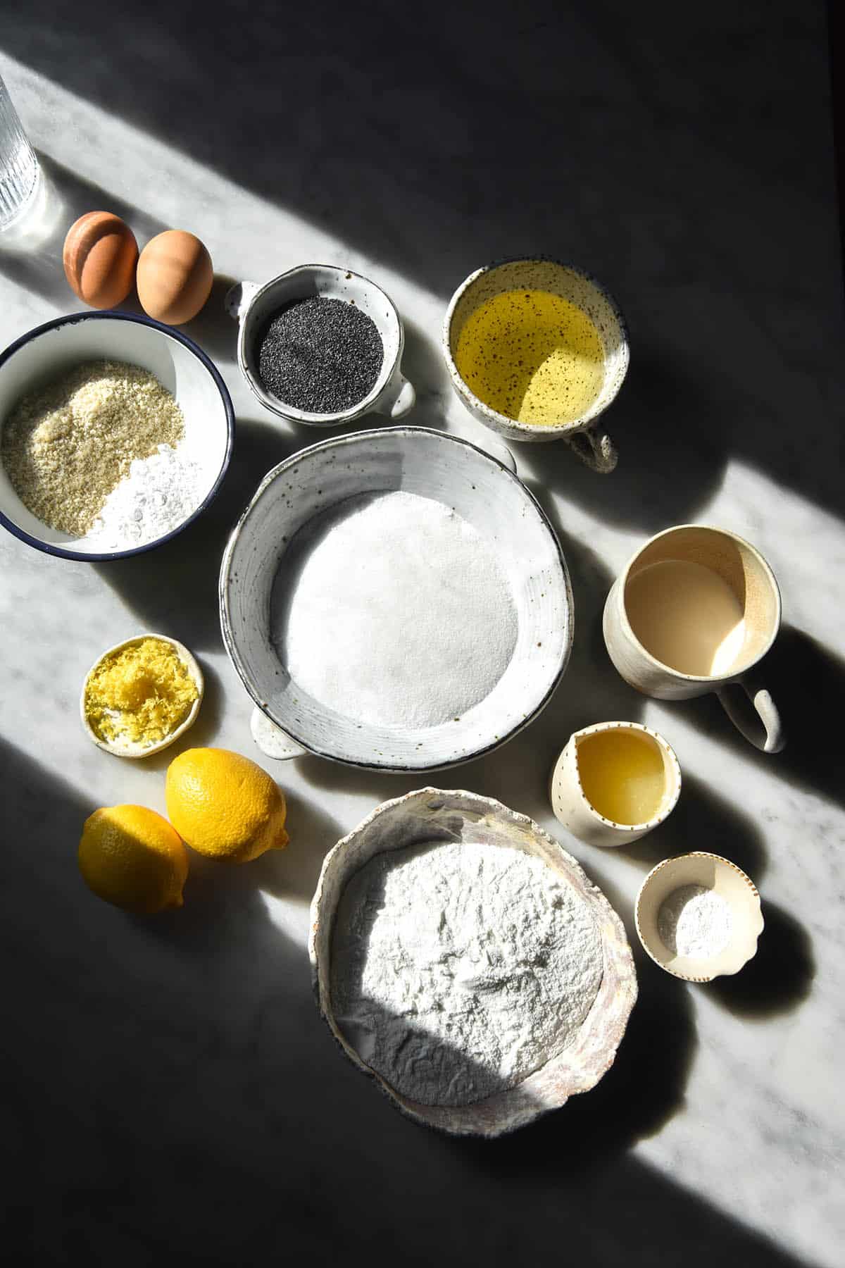 An aerial view of the ingredients used to make a gluten free lemon poppy seed cake in small white ceramic bowls on a white marble table. The ingredients are grouped together in a section of the table that is lit with bright sunlight. The rest of the image is in the shadow. 