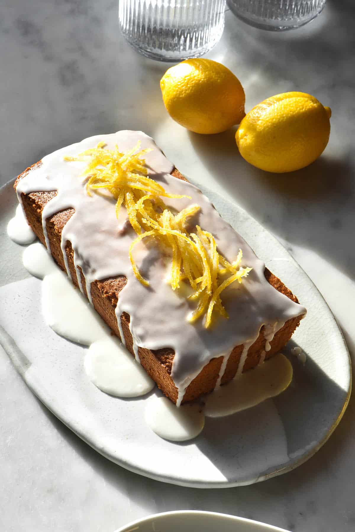 A sunlit image of a gluten free lemon poppy seed cake on a white and grey ceramic oval plate on a white marble table. The image is bathed in contrasting sunlight which accentuates the white lemon icing and candied lemon atop the loaf. 