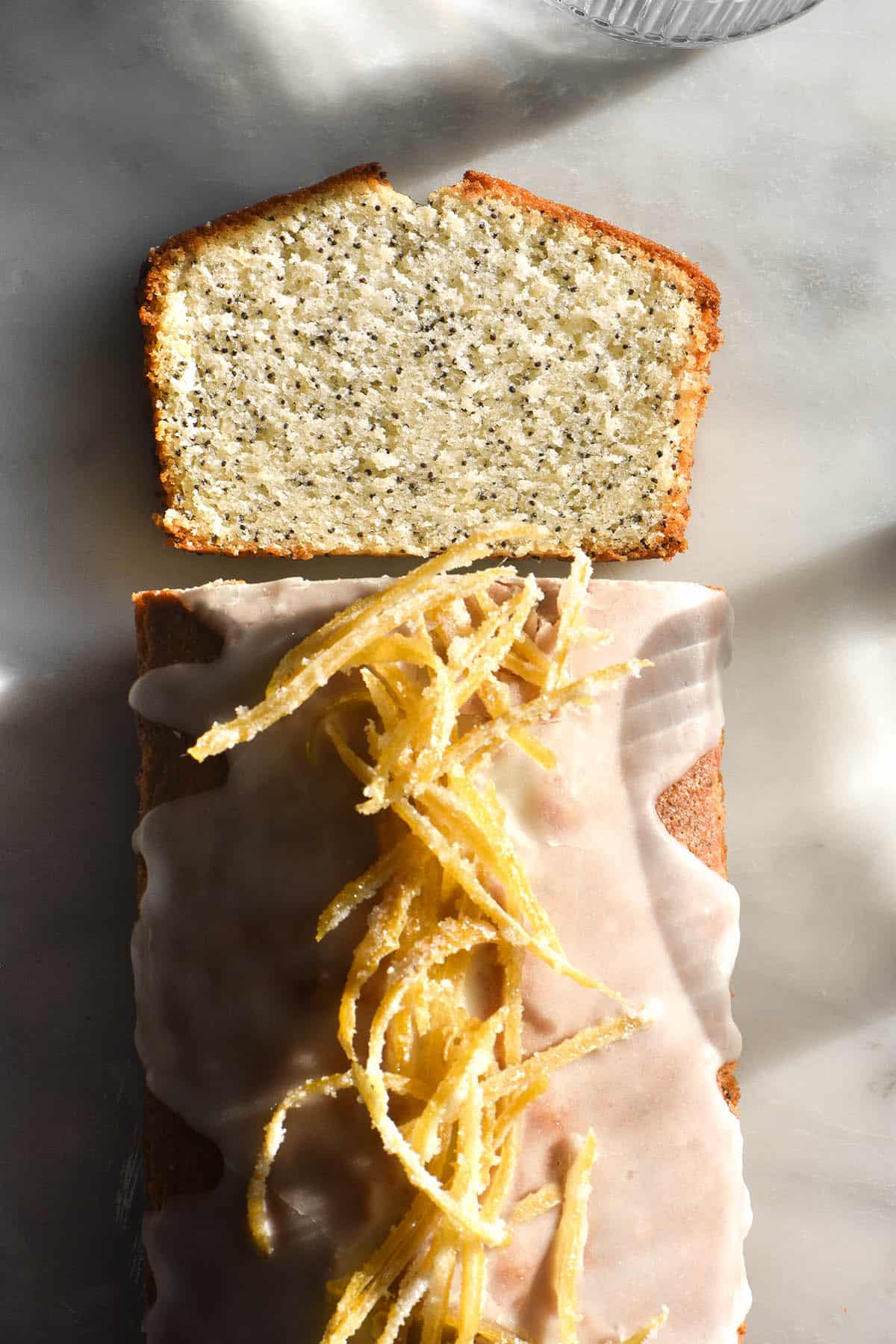 An aerial sunlit image of a gluten free lemon poppyseed loaf topped with white icing icing and candied lemon. A slice of the loaf lays on the table facing the camera, revealing the light and fluffy poppyseed crumb. 