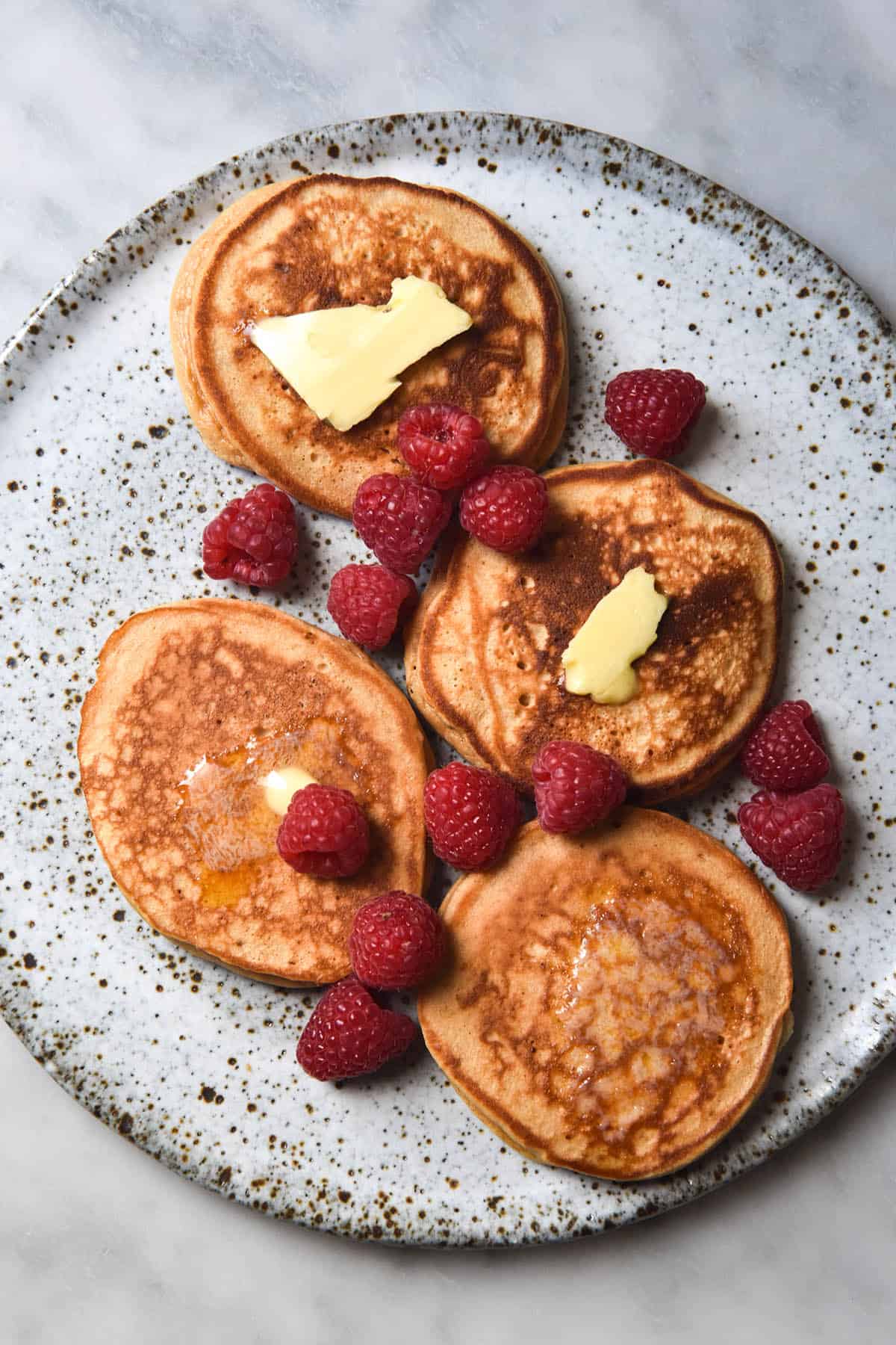 An aerial view of four gluten free protein pancakes on a white speckled ceramic plate. The pancakes are topped with melting chunks of butter and garnished with fresh raspberries.