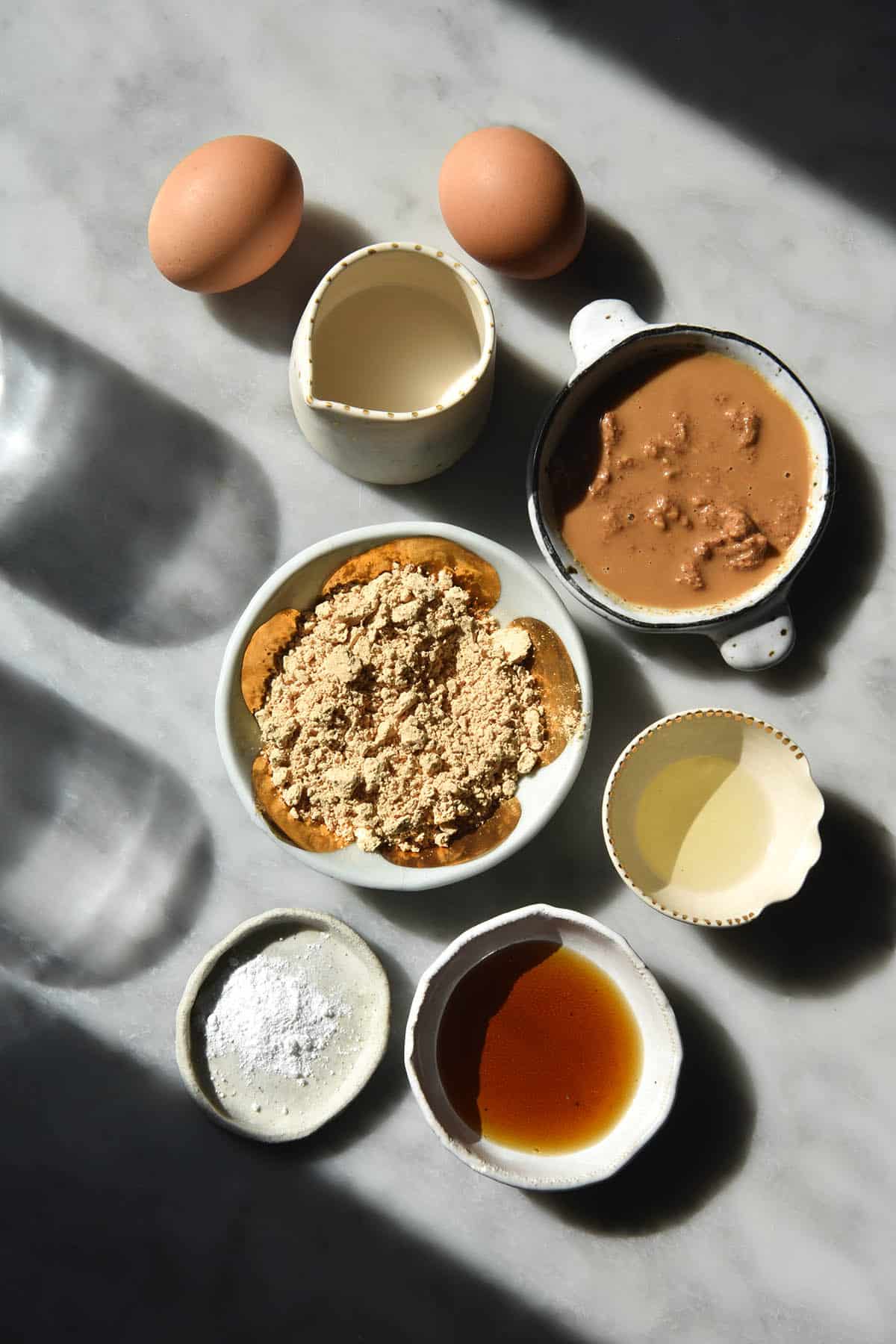 An aerial view of the ingredients for gluten free protein pancakes without banana on a white marble table in small ceramic bowls.