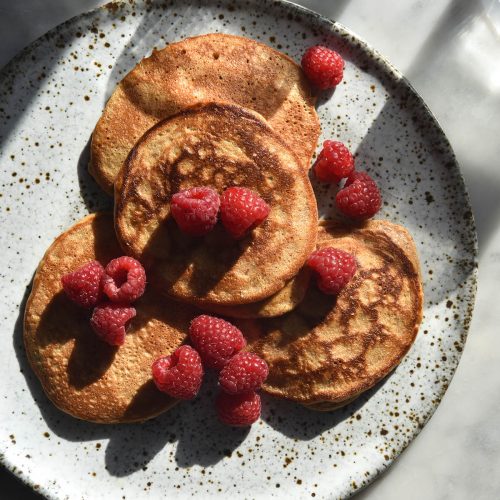 An aerial view of gluten free pancakes topped with fresh raspberries on a white speckled ceramic plate. Two water glasses sit in the top left corner and cast a shadow across the pancakes
