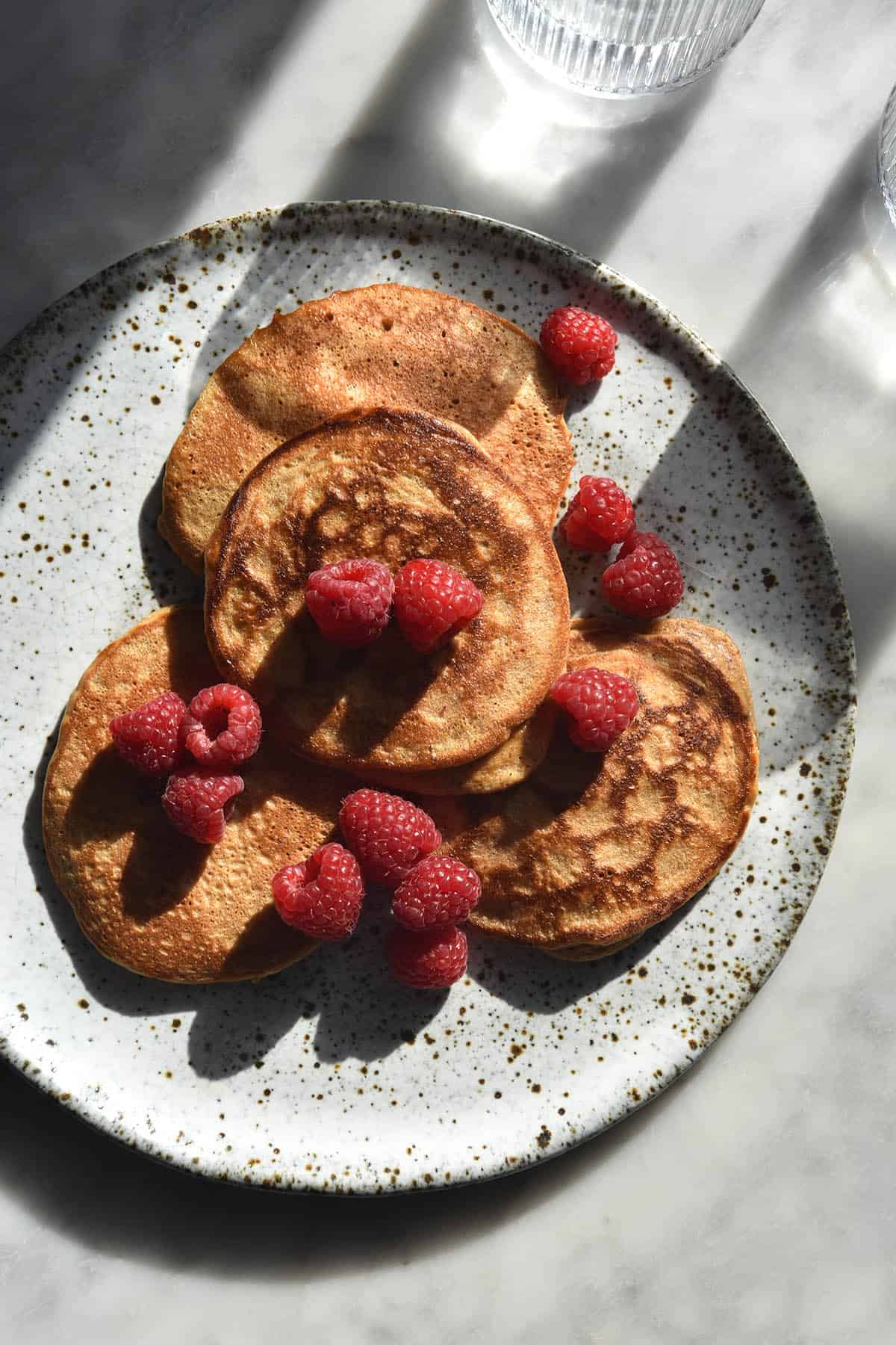 An aerial sunlit view of gluten free protein pancakes on a white ceramic plate topped with fresh raspberries. Two glasses of water sit in the top of the image and cast a shadow across the pancakes