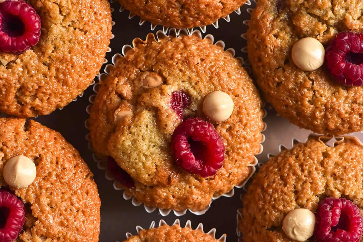 An aerial close up of a pale pink ceramic plate topped with gluten free raspberry muffins. The muffins are golden brown and studded with fresh raspberries and white chocolate chips. 