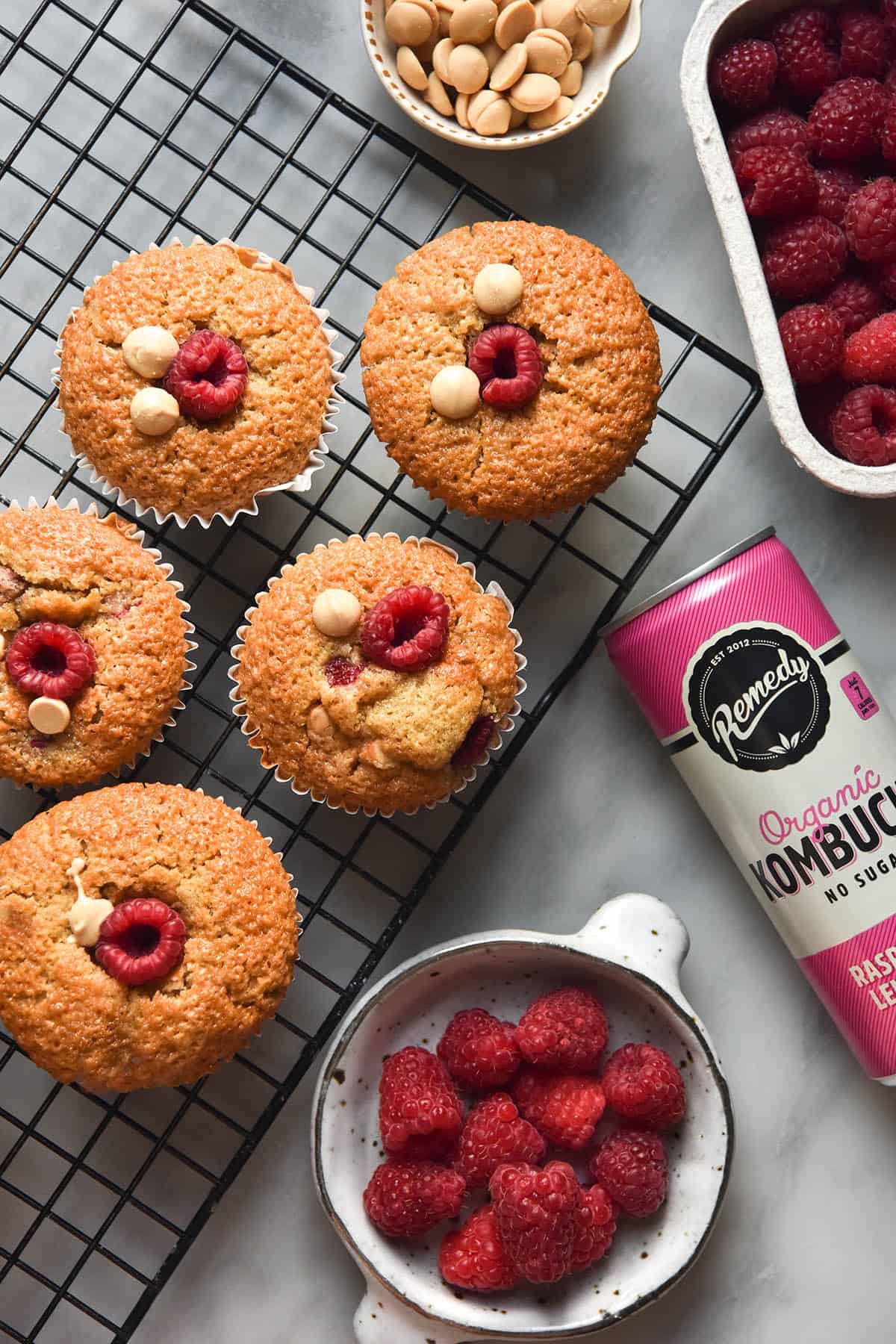 An aerial image of gluten free raspberry muffins on a baking rack atop a white marble table. The baking rack is surrounded by raspberries, kombucha and white chocolate chips, used in the muffins.