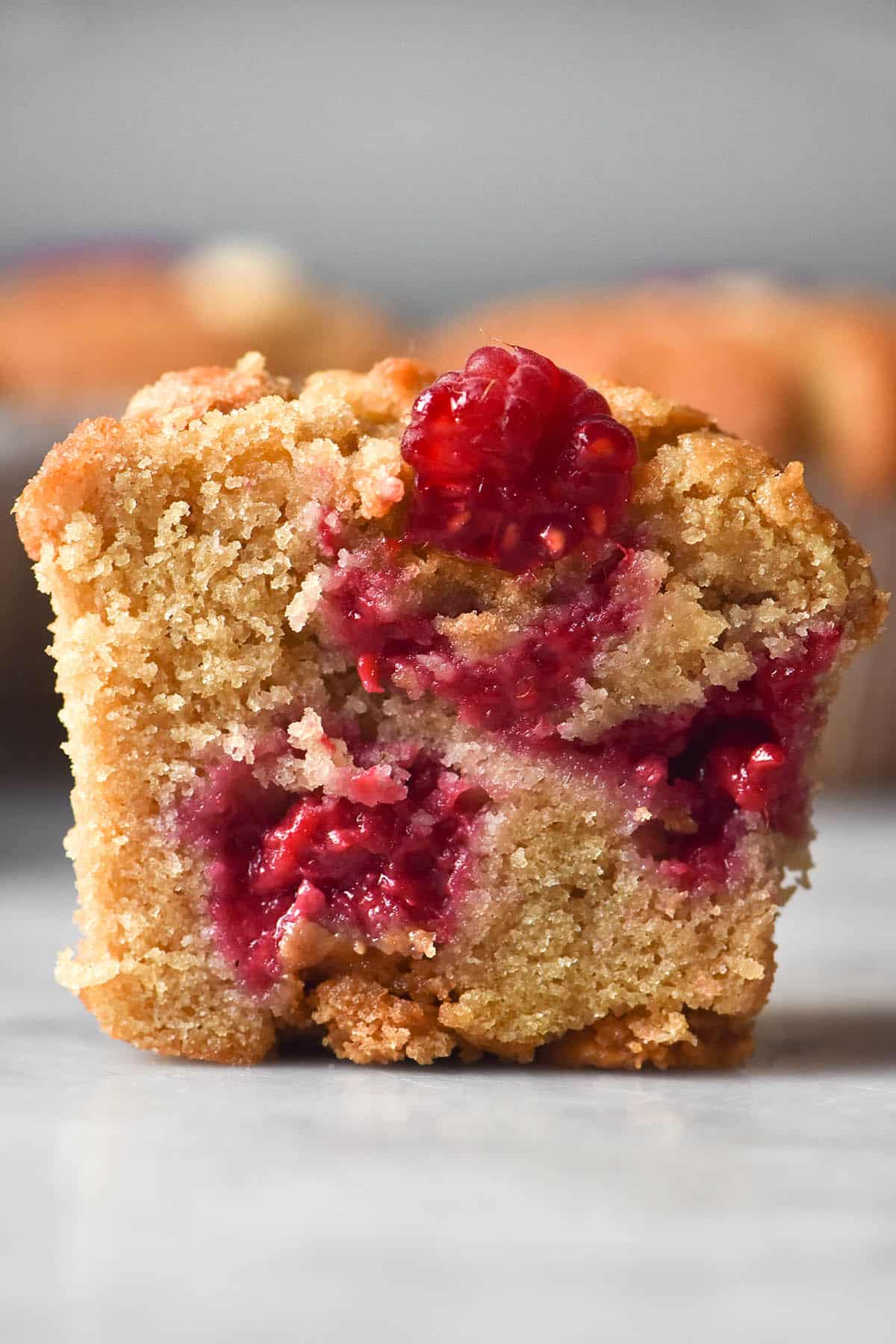 A side on macro image of a gluten free raspberry muffin sliced in half to reveal the raspberry stuffed crumb. The muffin sits on a white marble table with additional muffins in the background.