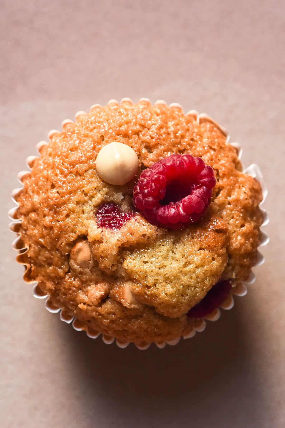 An aerial image of a gluten free chocolate chip muffin with raspberries. The muffin sits atop a pale pink ceramic plate