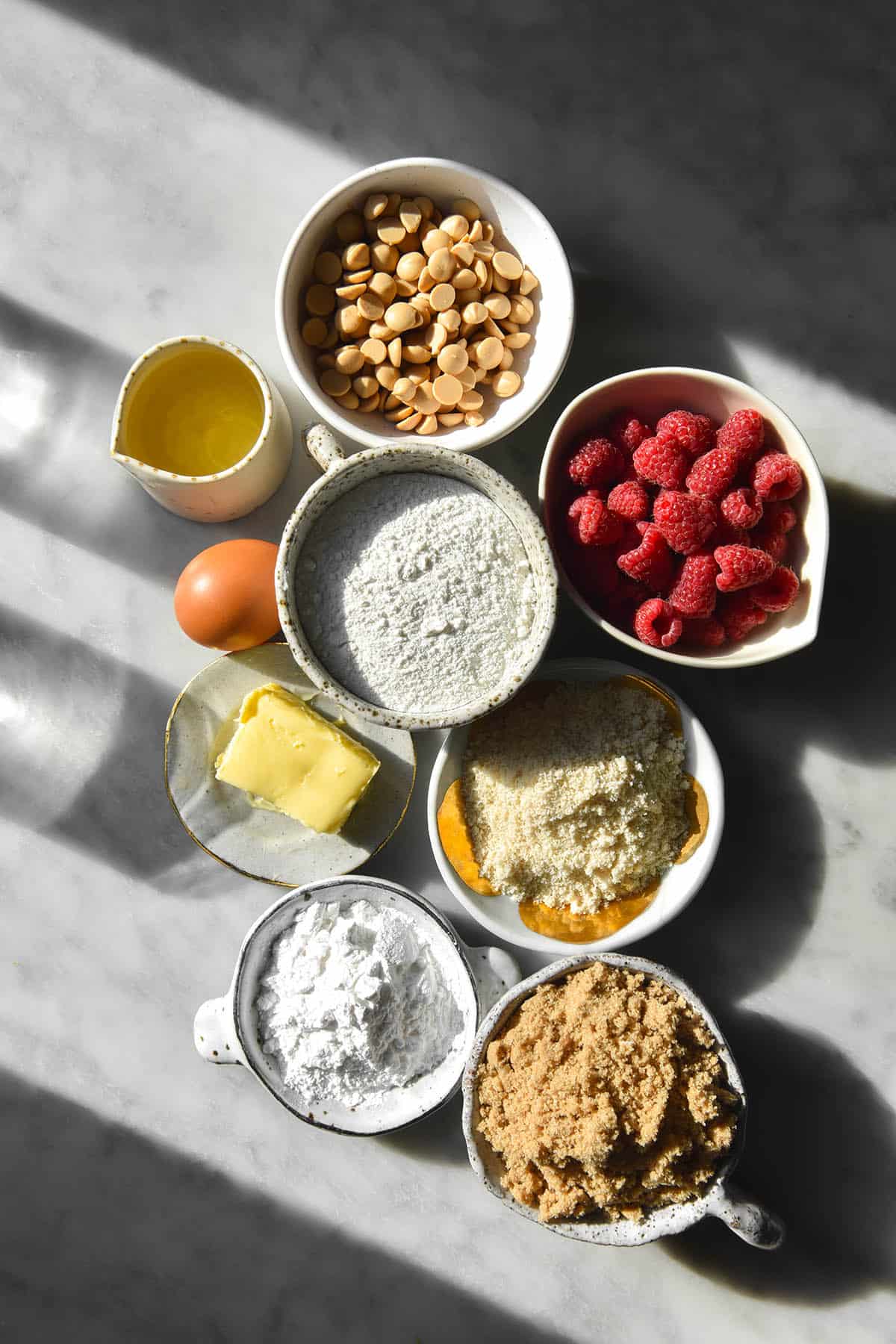 A moody contrasting sunlight image of the ingredients used for gluten free raspberry muffins on a white marble table. The ingredients are arranged in the centre of the table in white marble pinch bowls and two glasses of water sit off to the left side