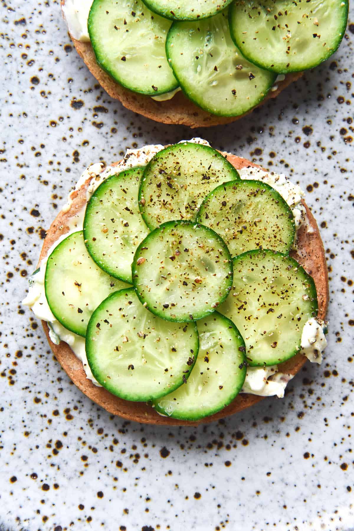 An aerial image of a gluten free vegan bagel sliced in half and topped with vegan cream cheese, thinly sliced cucumber, salt and pepper. The slices sit atop a white speckled ceramic plate. 