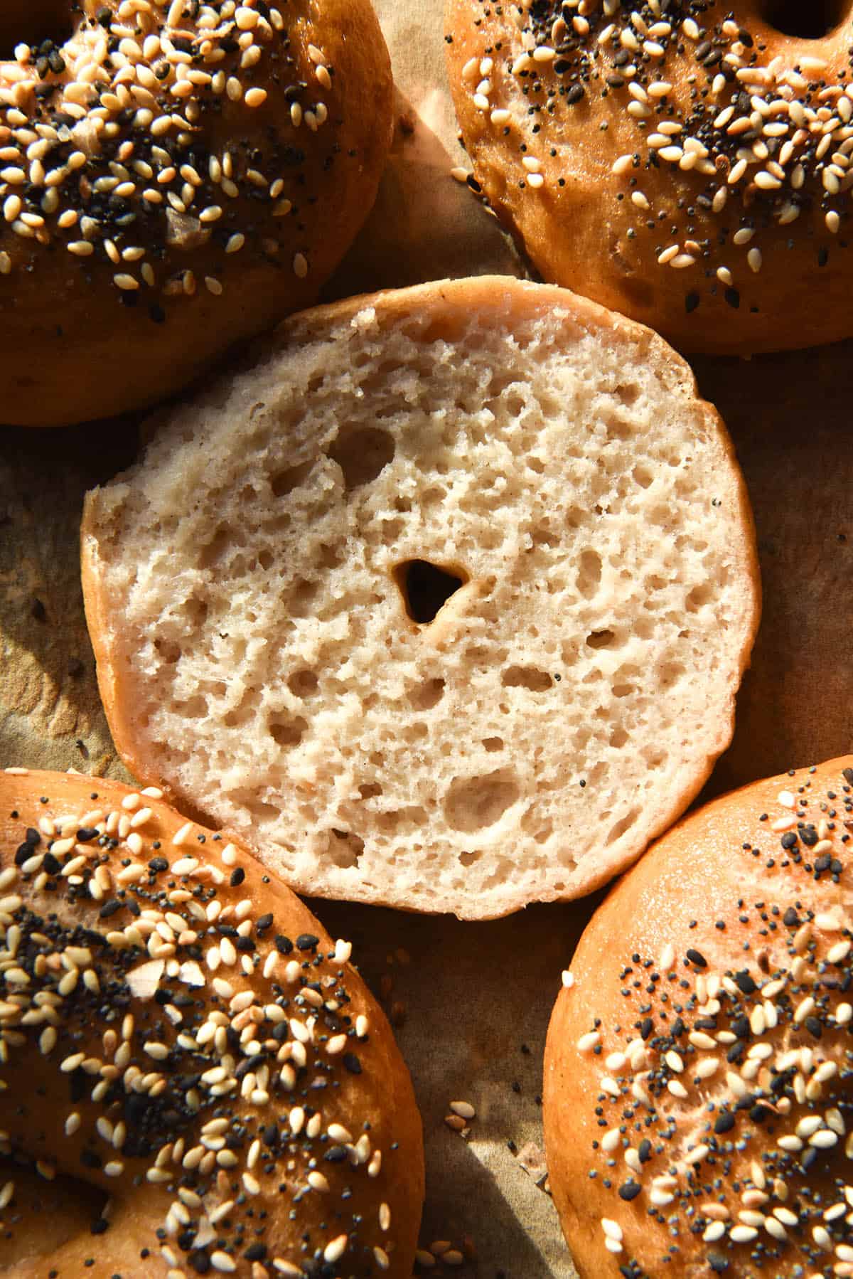 An aerial close up of the crumb of a sliced gluten free vegan bagel surrounded by other bagels topped with everything bagel seasoning on a baking sheet
