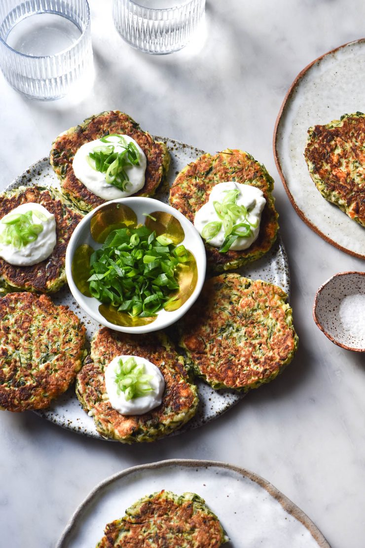 An aerial view of a a white speckled ceramic plate topped with a circle of gluten free zucchini fritters topped with a dollop of yoghurt and spring onion greens. The fritters are in a circle surrounding a small bowl filled with extra spring onion greens