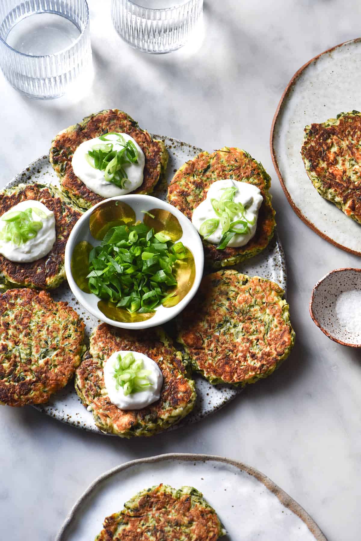 An aerial image of gluten free zucchini fritters on a white speckled ceramic plate atop a white marble table. Some of the fritters are topped with yoghurt and spring onion greens. A small bowl of spring onion greens sits in the centre of the plate, which is surrounded by extra plates of fritters, water glasses and a bit bowl of salt.