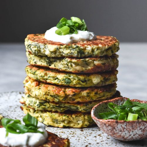 A side on view of a stack of gluten free zucchini fritters on a white speckled ceramic plate atop a white marble table. The top fritter is topped with yoghurt and some spring onion greens, and a pinch bowl of extra greens sits to the right of the stack. Another fritter sits in the front left of the image.