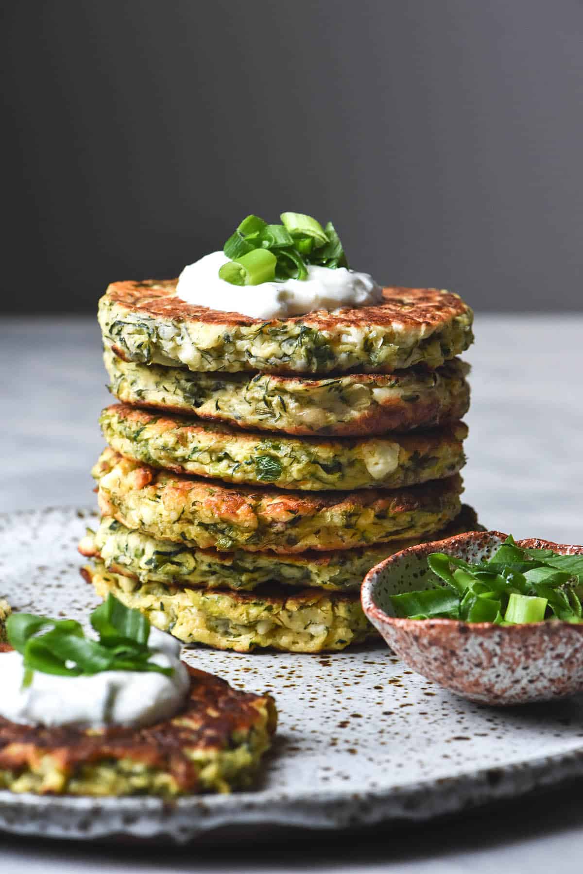 A side on view of a stack of gluten free zucchini fritters on a white speckled ceramic plate atop a white marble table. The top fritter is topped with yoghurt and some spring onion greens, and a pinch bowl of extra greens sits to the right of the stack. Another fritter sits in the front left of the image.