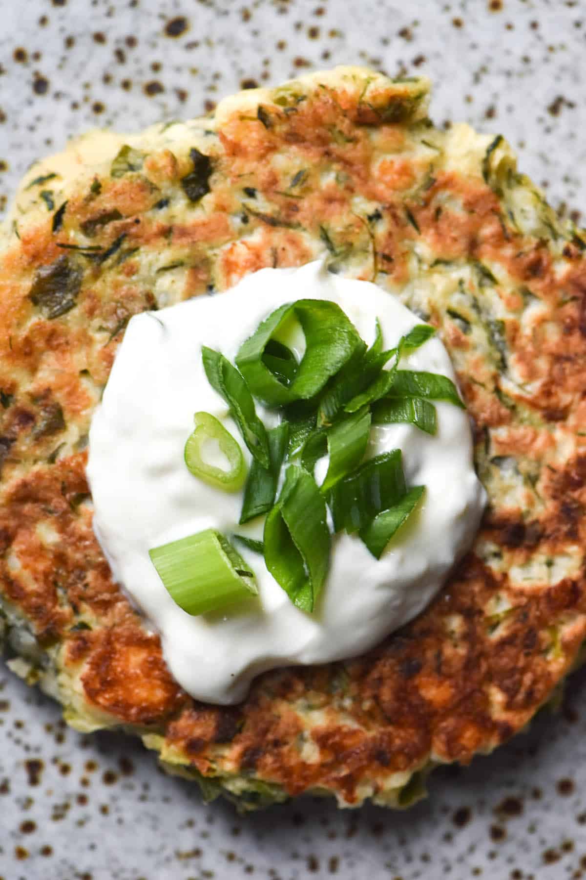 An aerial close up view of a gluten free zucchini fritter topped with a dollop of yoghurt and spring onion greens on a white speckled ceramic plate 