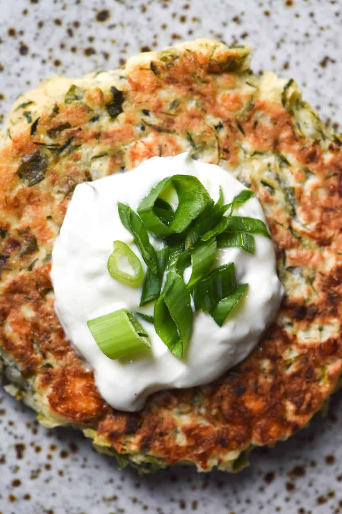 An aerial close up view of a gluten free zucchini fritter topped with a dollop of yoghurt and spring onion greens on a white speckled ceramic plate