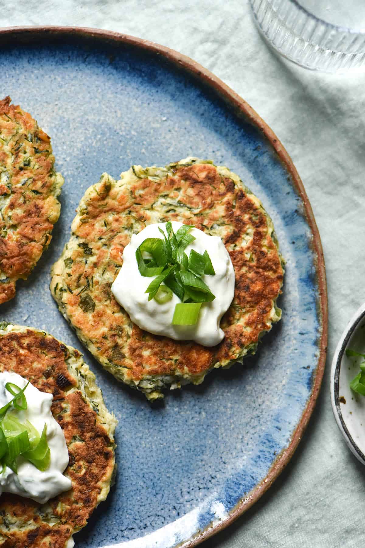An aerial view of three gluten free zucchini fritters topped with a dollop of yoghurt and spring onion greens on a bright blue ceramic plate. The plate sits on a pale green linen tablecloth and a glass of water and pinch bowl sit to the right of the plate