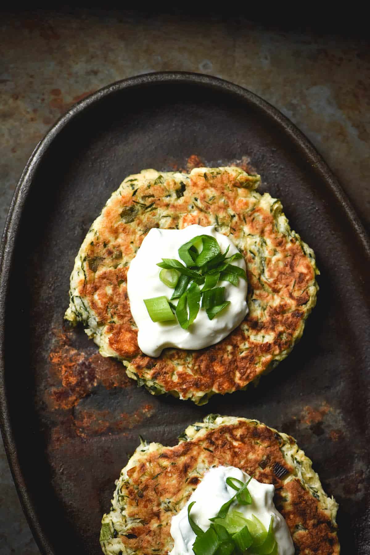 An aerial image of two gluten free zucchini fritters topped with a dollop of yoghurt and spring onion greens on a lightly rusted sizzle platter against a dark metal backdrop.