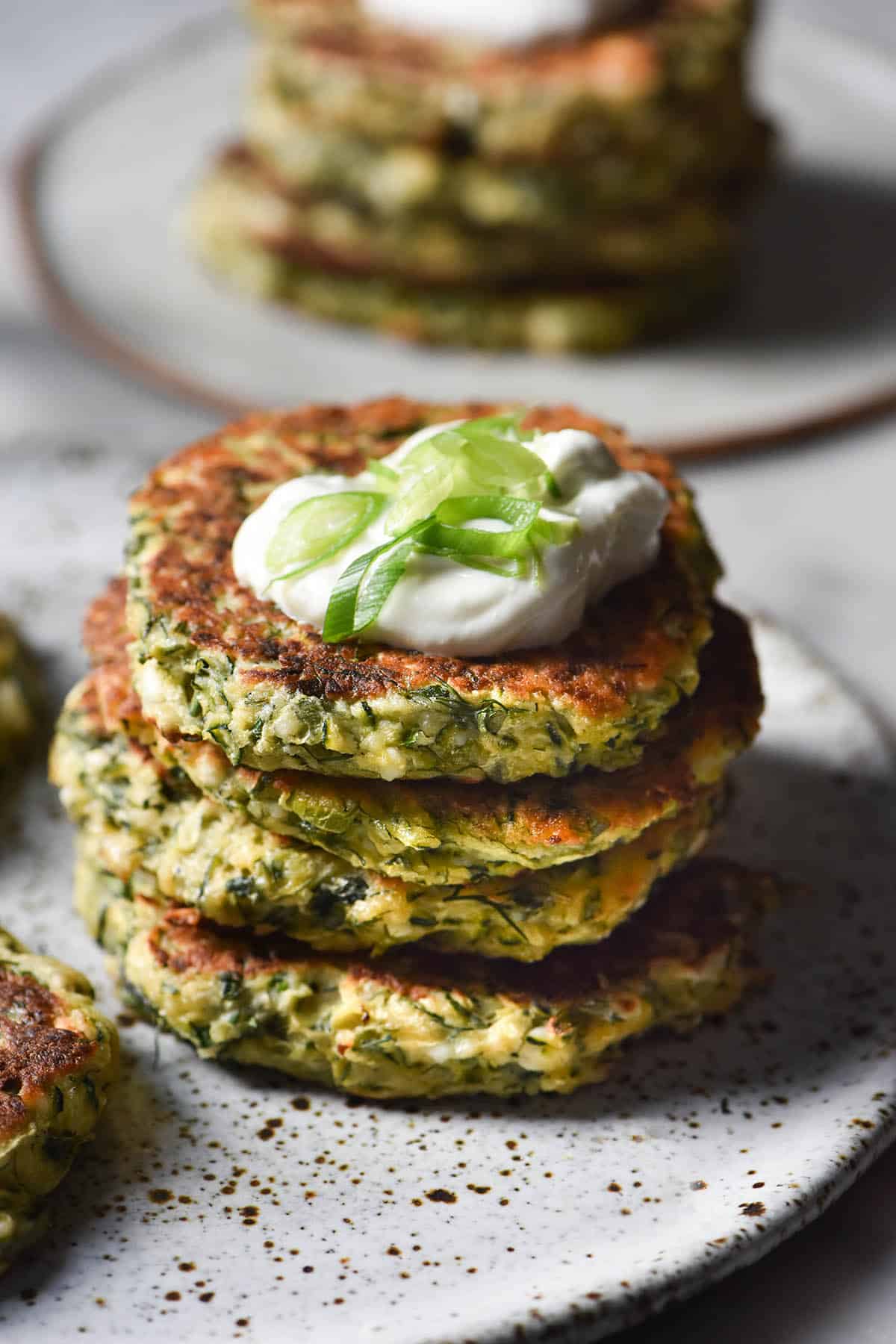 A side on view of a stack of gluten free zucchini fritters on a white speckled ceramic plate atop a white marble table. The stack is topped with a dollop of yoghurt and spring onion greens, and a second stack on a white plate sits in the background