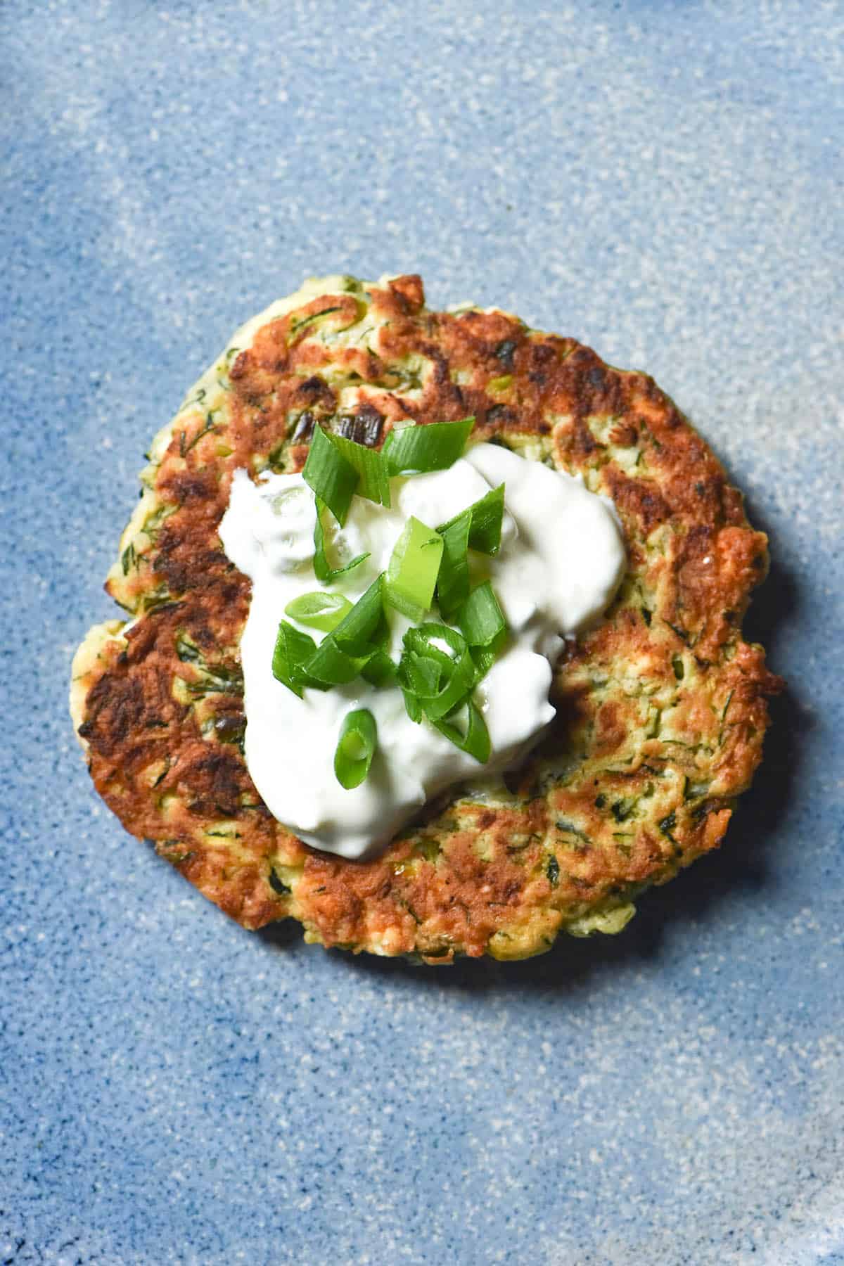 An aerial view of a gluten free zucchini fritter topped with a dollop of yoghurt and spring onion greens atop a bright blue ceramic plate