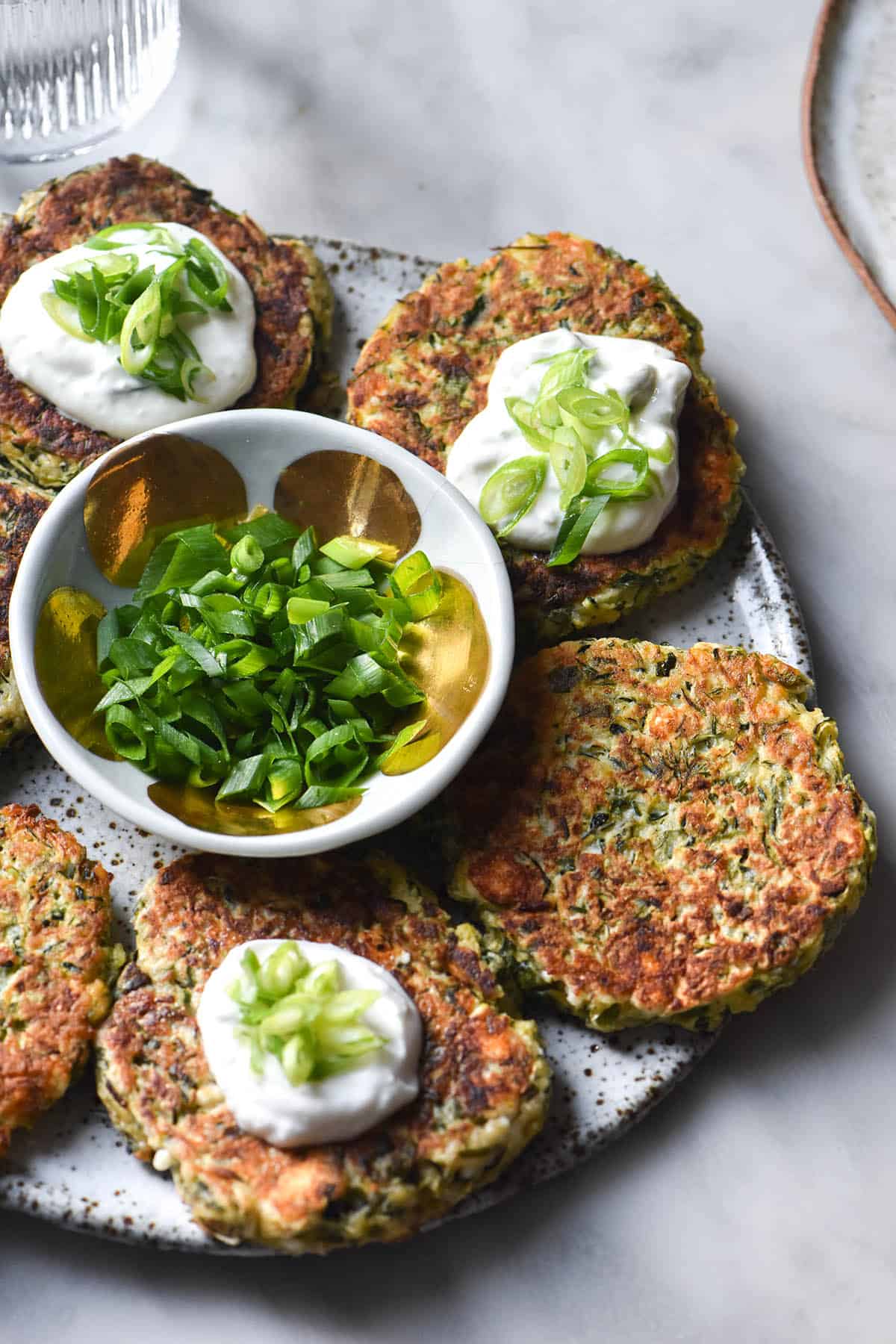 An aerial view of gluten free zucchini fritters topped with a dollop of yoghurt and spring onion greens on a white speckled ceramic plate. The fritters are in a circle surrounding a small bowl filled with extra spring onion greens. A glass of water and an extra white plate sit on the white marble table background