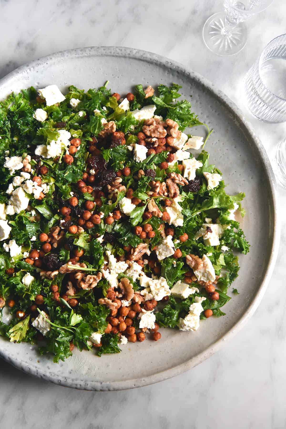 An aerial image of a kale, feta, raisin and walnut salad in a large white ceramic serving dish. The dish sits atop a white marble table and is surrounded by water glasses. 