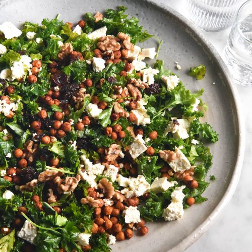 An aerial image of a kale, feta, raisin and walnut salad in a large white ceramic serving dish. The dish sits atop a white marble table and is surrounded by water glasses.