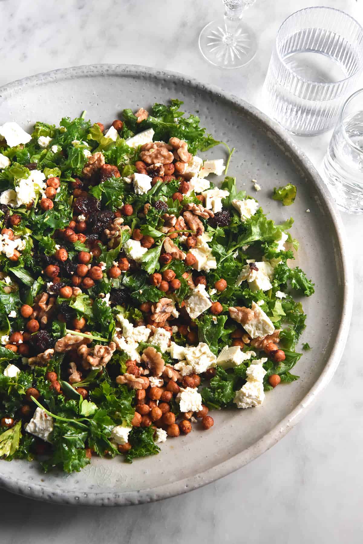 An aerial image of a kale, feta, raisin and walnut salad in a large white ceramic serving dish. The dish sits atop a white marble table and is surrounded by water glasses.