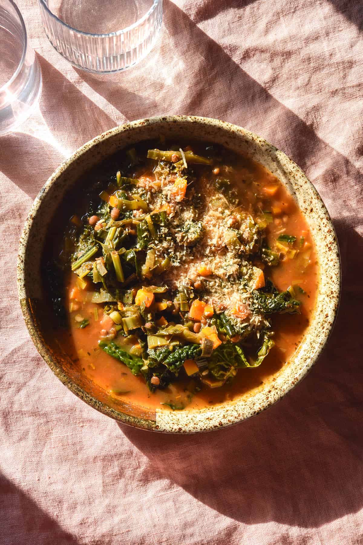 A sunlit aerial image of a bowl of low FODMAP lentil soup in a speckled beige bowl atop a pale pink linen tablecloth. The soup has a deep red broth and is topped with freshly grated parmesan 