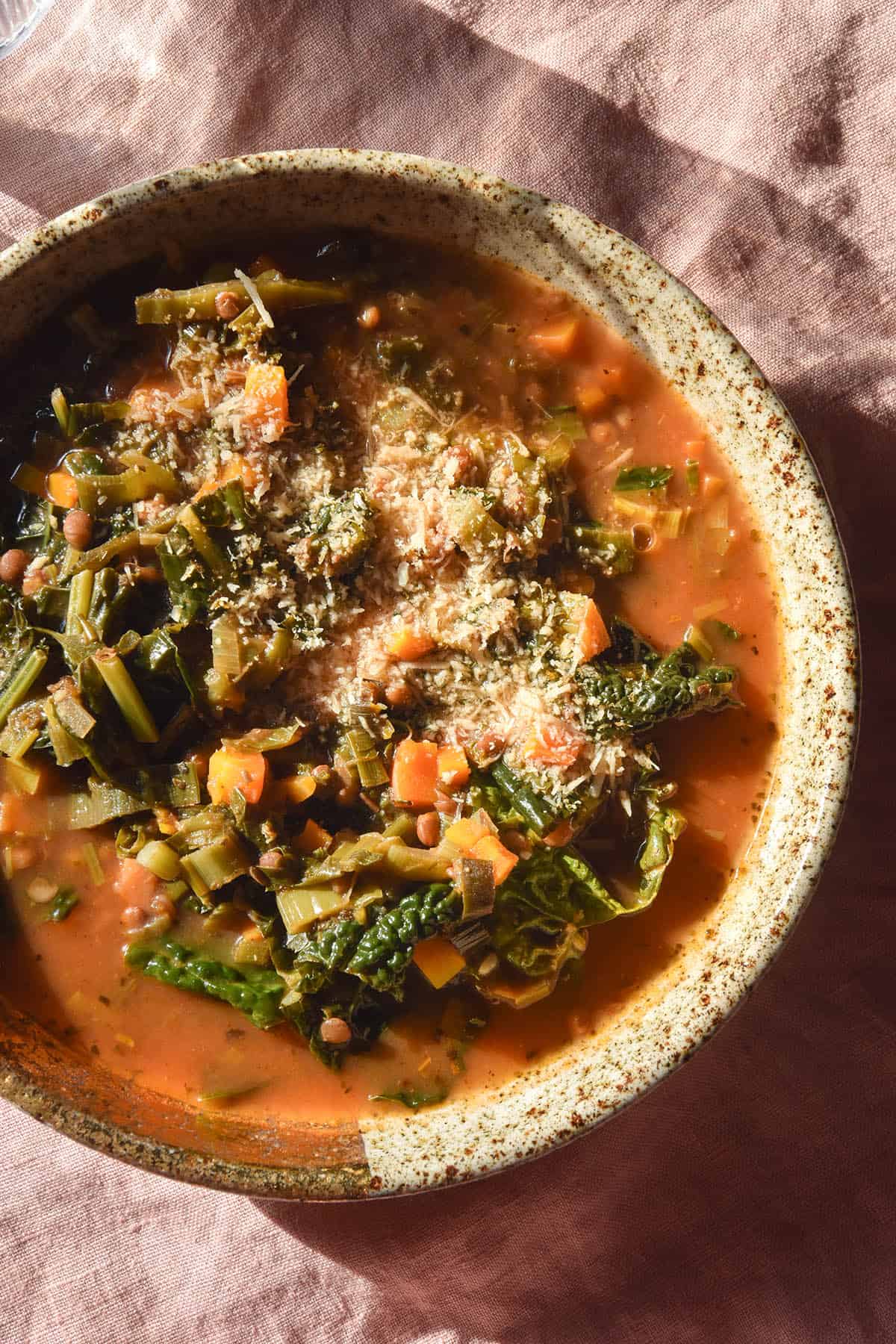 A sunlit aerial image of a bowl of low FODMAP lentil soup in a speckled beige bowl atop a pale pink linen tablecloth. The soup has a deep red broth and is topped with freshly grated parmesan 