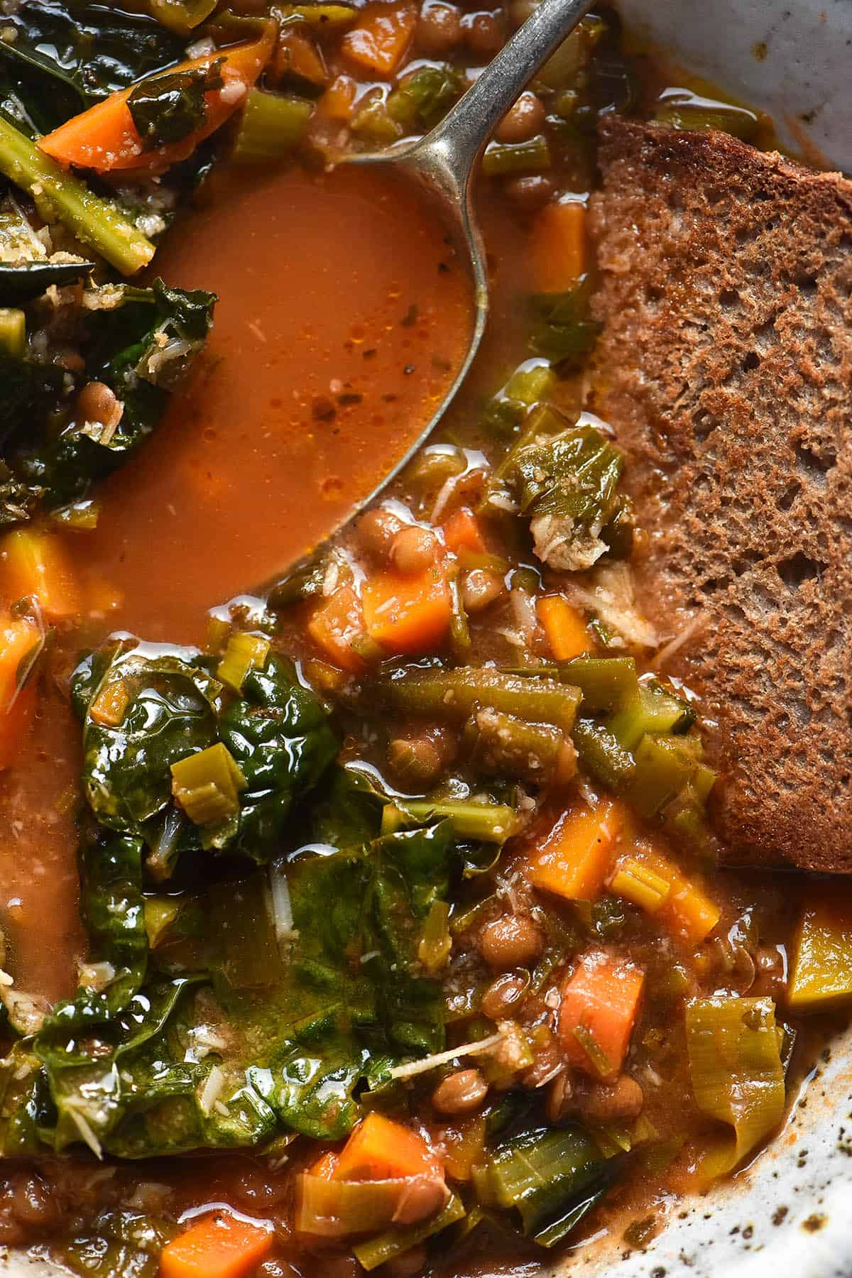 A close up macro view of a bowl of low FODMAP lentil soup with buckwheat toast dipping into the top right corner. A spoon sits in the top left corner, revealing the bright red broth of the soup. 