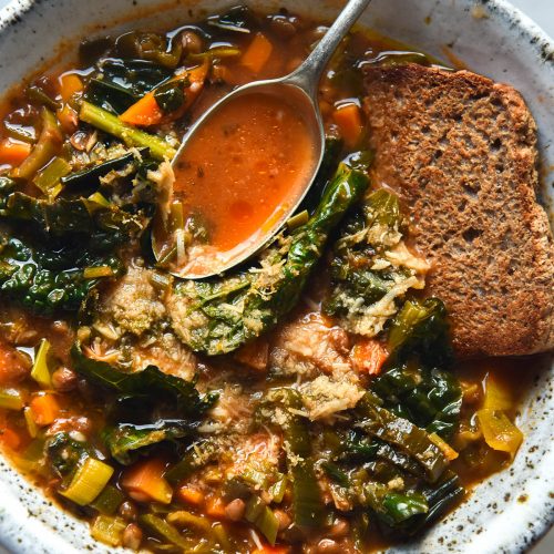 An aerial close up view of a bowl of low FODMAP lentil soup topped with grated parmesan and served with a slice of buckwheat bread. The soup sits in a white speckled ceramic bowl atop a white marble table.