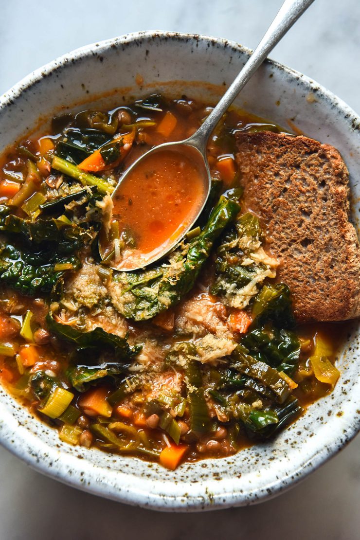 An aerial close up view of a bowl of low FODMAP lentil soup topped with grated parmesan and served with a slice of buckwheat bread. The soup sits in a white speckled ceramic bowl atop a white marble table.