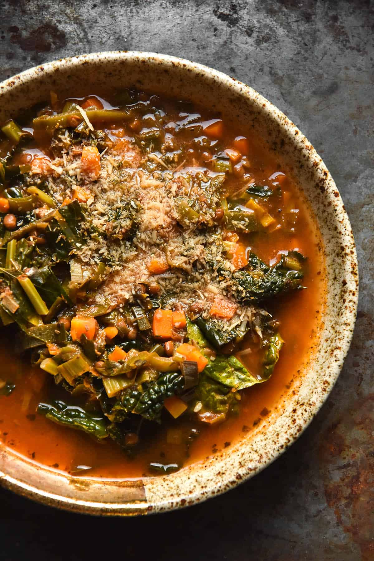 An aerial close up view of a bowl of low FODMAP lentil soup topped with finely grated parmesan. The soup sits in a beige speckled ceramic bowl atop a grey steel backdrop. 