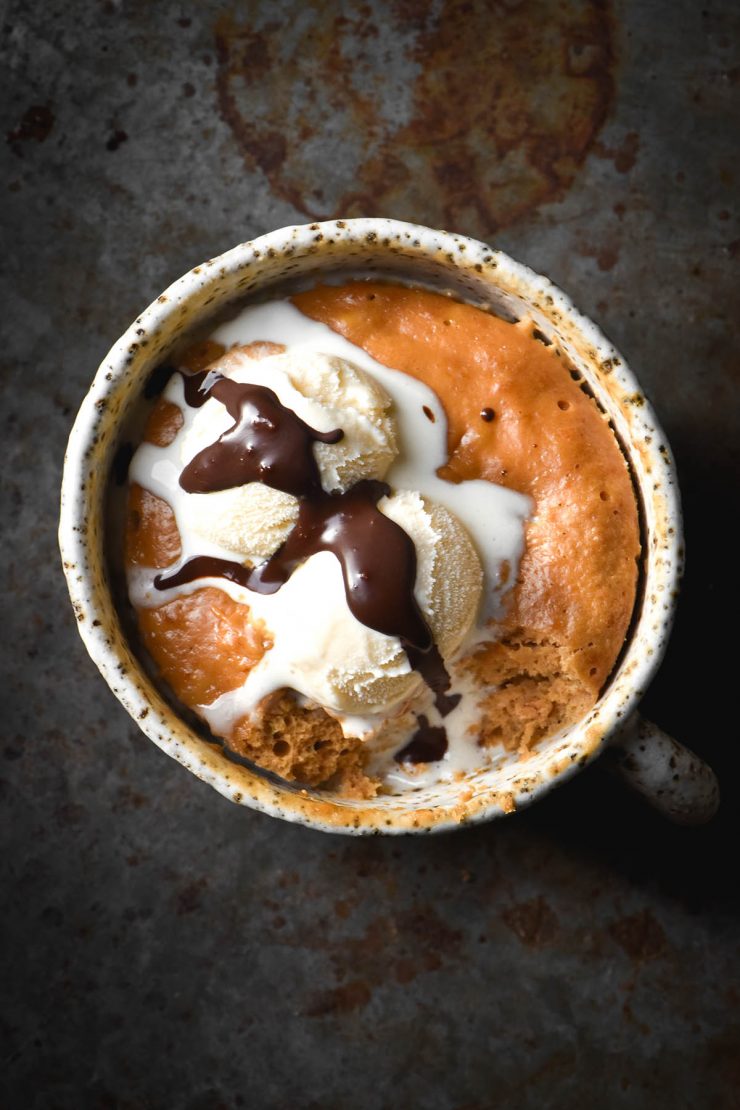 An aerial view of a protein mug cake topped with melting vanilla ice cream and melted chocolate. The cake sits in a white speckled ceramic mug which sits on a dark metal steel backdrop