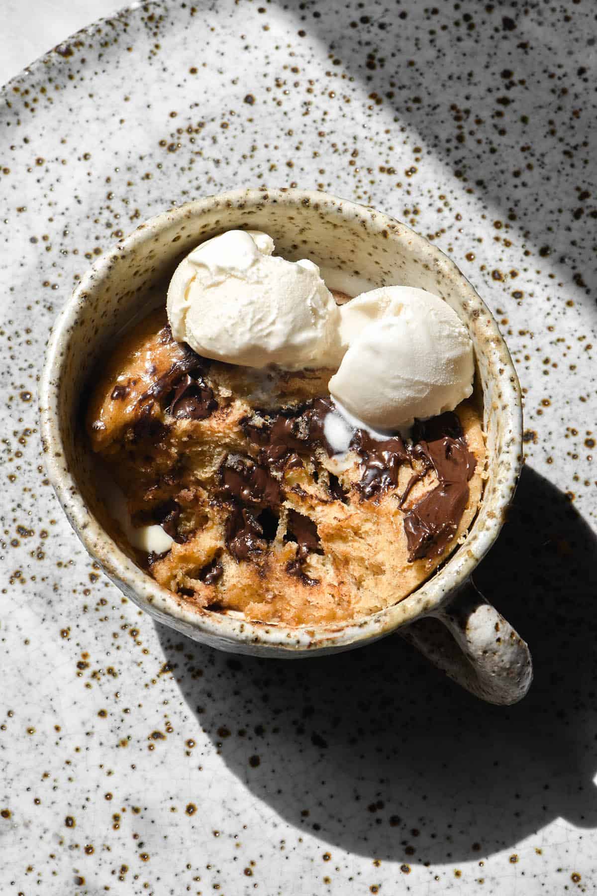 A sunlit aerial image of a protein mug cake with chocolate chips and a small scoop of vanilla ice cream. The mug cake sits in a white speckled mug atop a white speckled ceramic plate. 