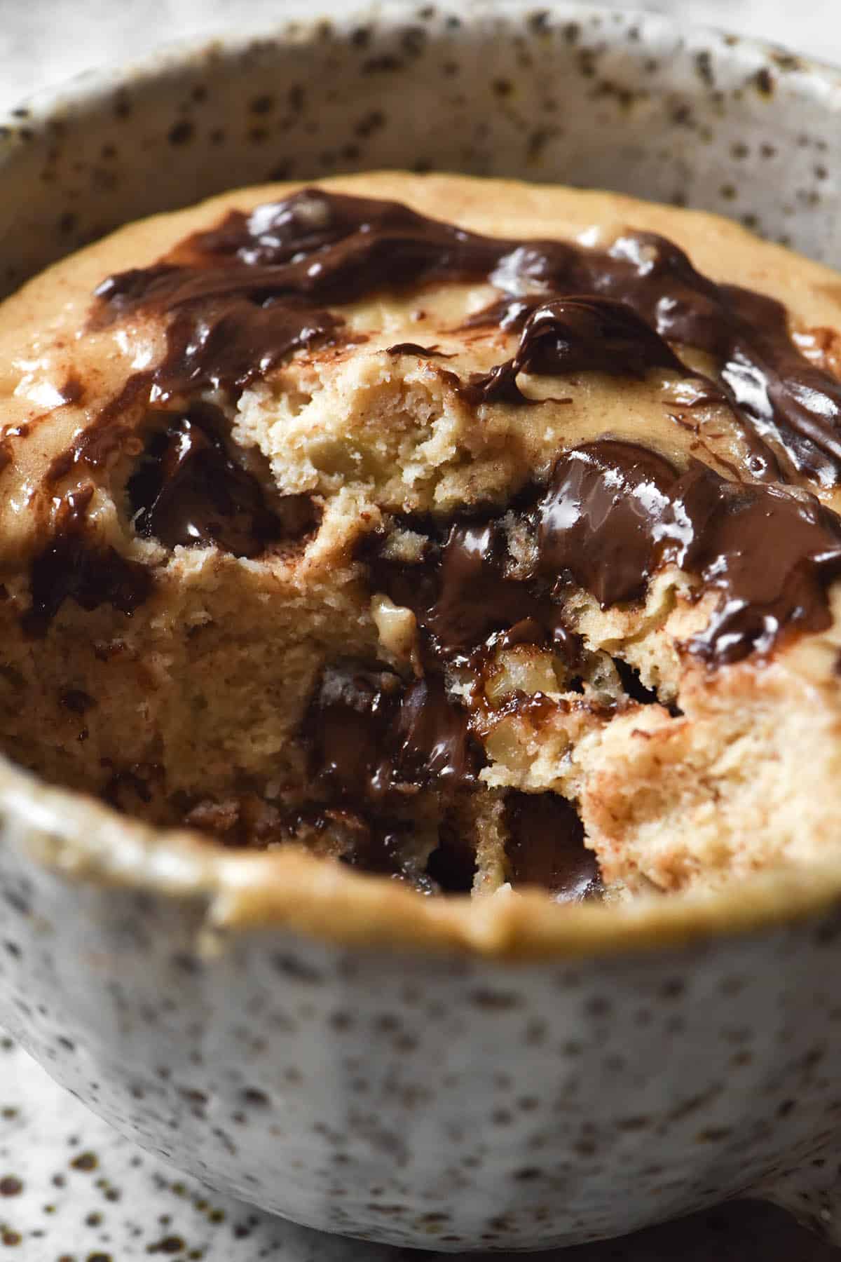 A side on macro close up image of a protein mug cake filled with melty chocolate chips. The front part of the mug cake has been eaten, revealing the melty chocolate and crumb within. The cake is in a white speckled ceramic mug. 